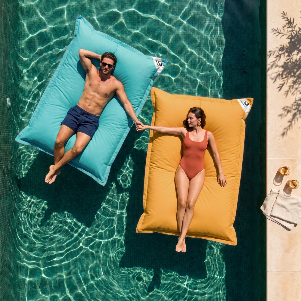 Solo funda, para rellenar – Dos pufs gigantes flotantes Deco Arts, Azul Caribe y Amarillo Miel, en una piscina con una pareja, vista cenital